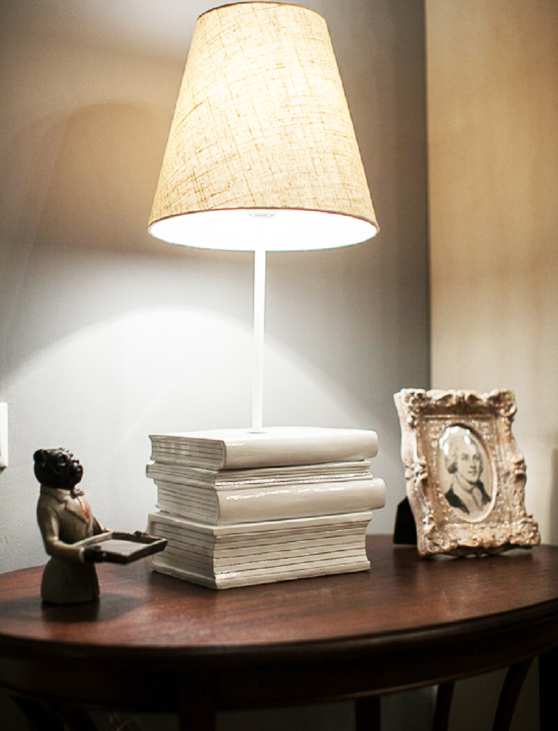 Het tafelblad van de ronde tafel is versierd met een ongewone tafelvaas met een linnen lampenkap, geïnstalleerd op een voetsteun in de vorm van boeken