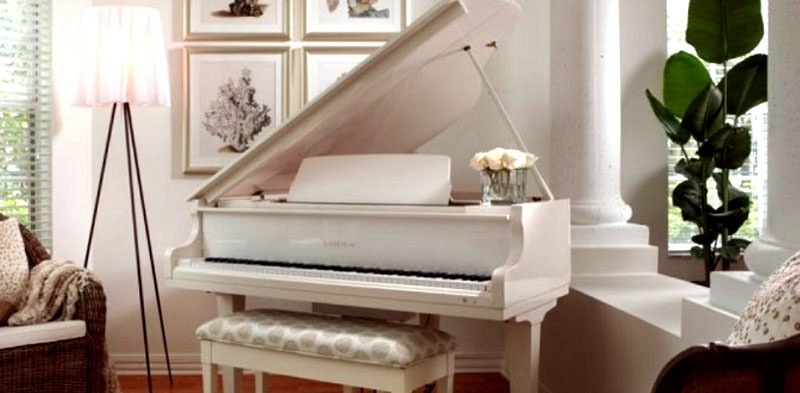 Przy fortepianie stoi designerska lampa podłogowa na trójnogu z kloszem z delikatnej prześwitującej tkaniny
